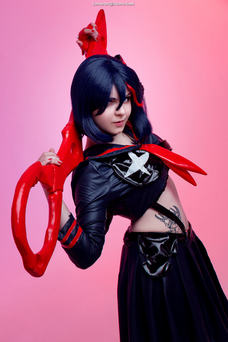 A person wearing a black and red costume with a large red weapon, posing against a pink background.