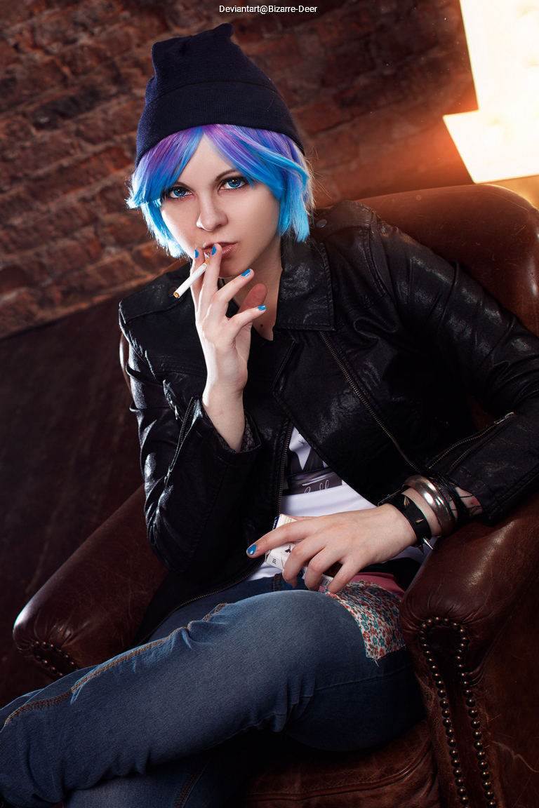 A person with short blue and purple hair wearing a black leather jacket and a beanie, seated on a brown leather armchair, holding a cigarette and looking confidently at the camera.