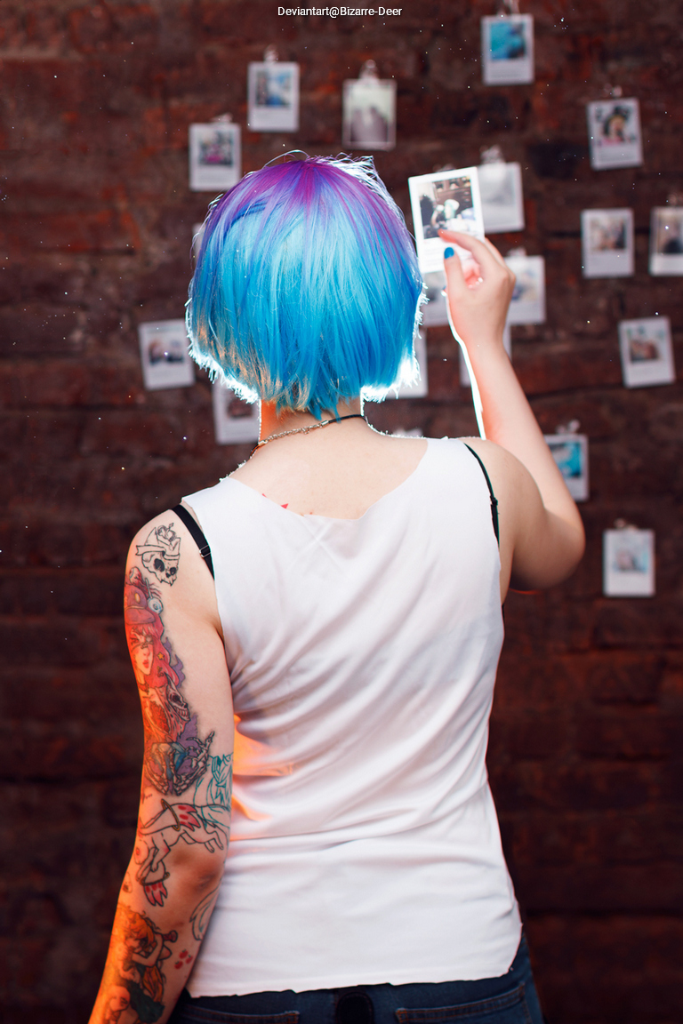A person with blue and purple hair stands with their back to the camera, holding a printed photo in front of a wall filled with several hanging photographs. They are wearing a white tank top and have colorful tattoos visible on their arms.