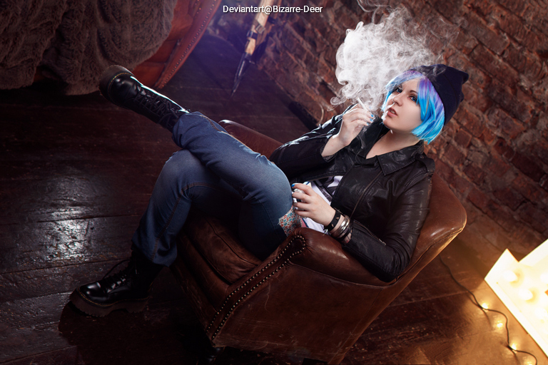 A person with blue hair wearing a black leather jacket sits in a vintage armchair, smoking and looking contemplative, with a backdrop of exposed brick walls.