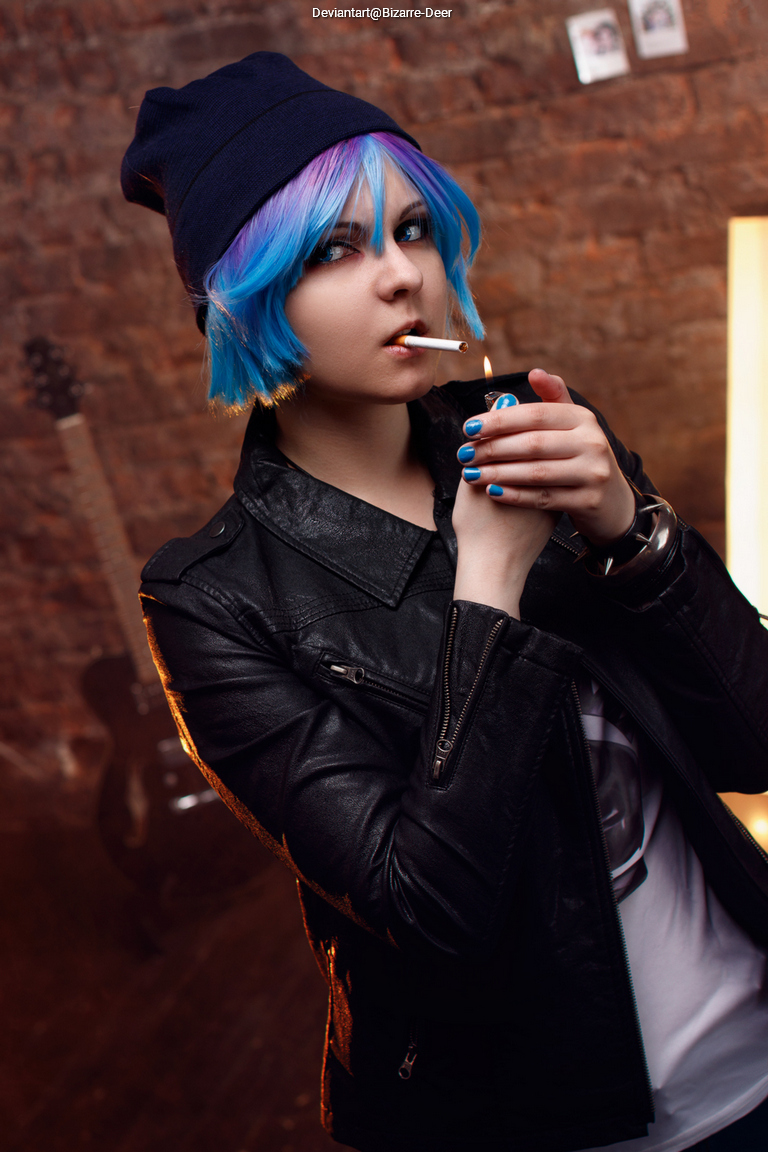 A person with short blue and purple hair wearing a black leather jacket and a beanie, holding a cigarette and a drink in a dimly lit room with brick walls.