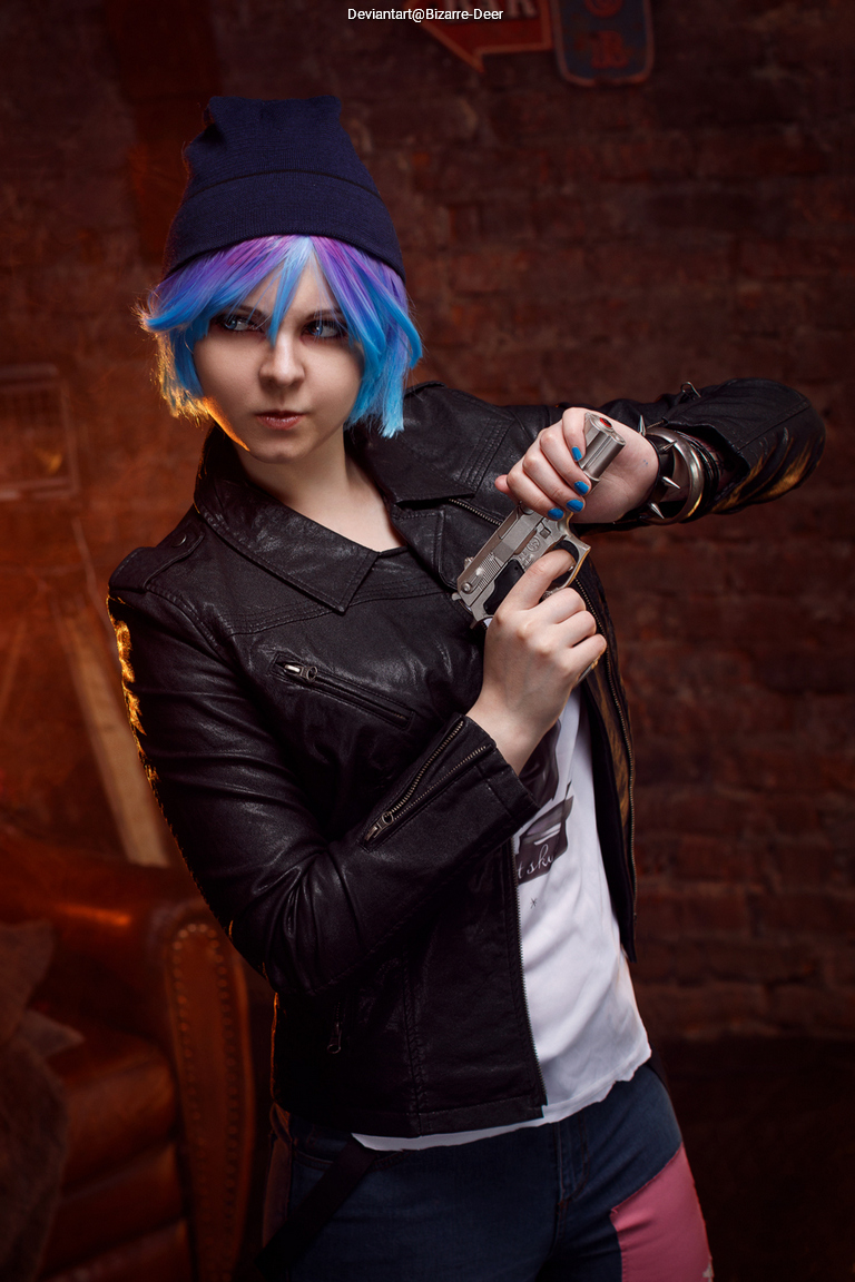 A person with colorful hair, wearing a black leather jacket and a beanie, poses confidently while holding a gun.