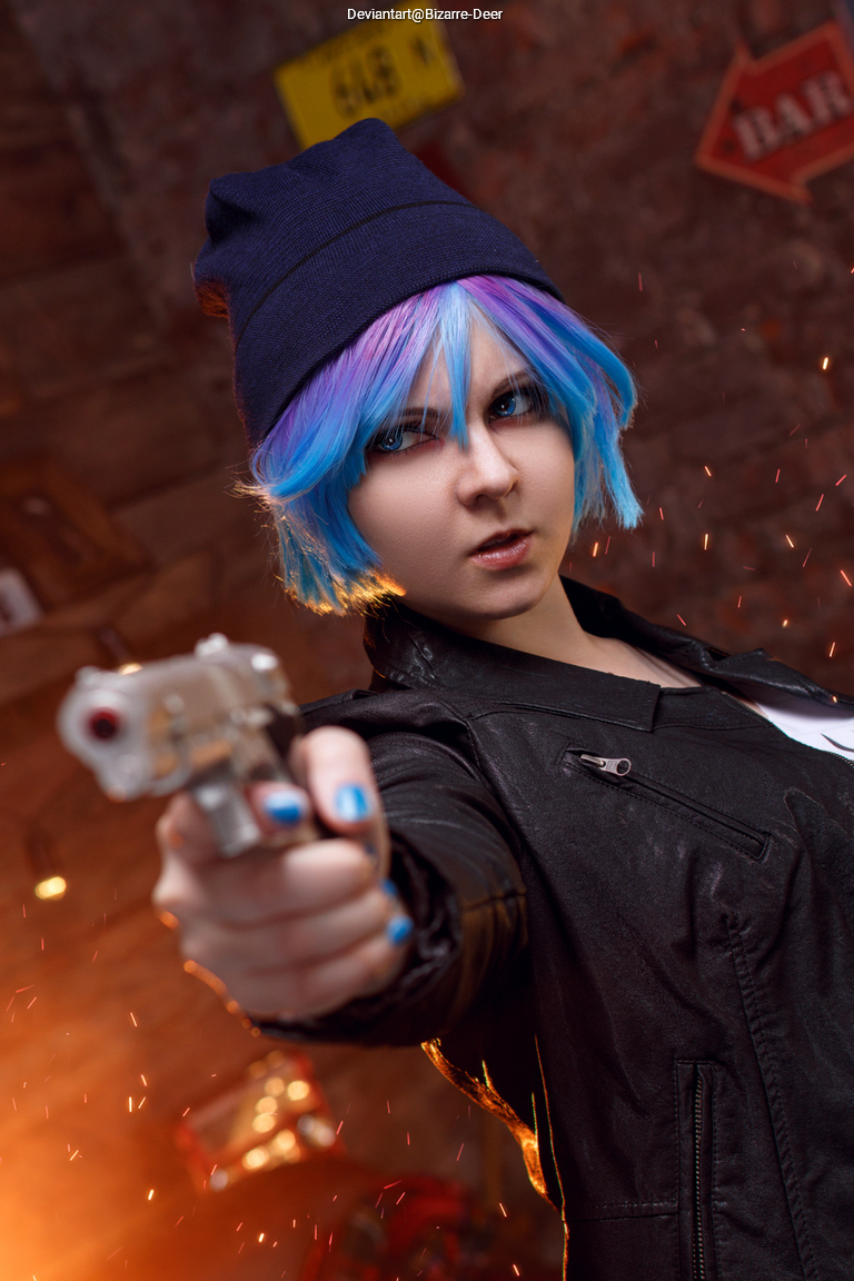 Person with colorful hair and a blue beanie holding a toy gun, dressed in a black leather jacket, posed dramatically against a brick wall with blurred lights in the background.