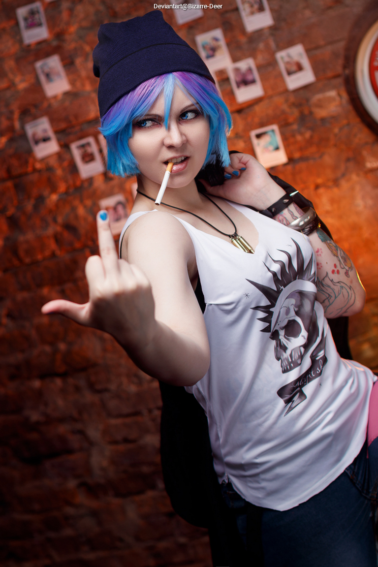 A person with blue and purple hair wearing a beanie and a sleeveless shirt poses in front of a rustic wall, showing a rebellious gesture with one hand while holding a cigarette.