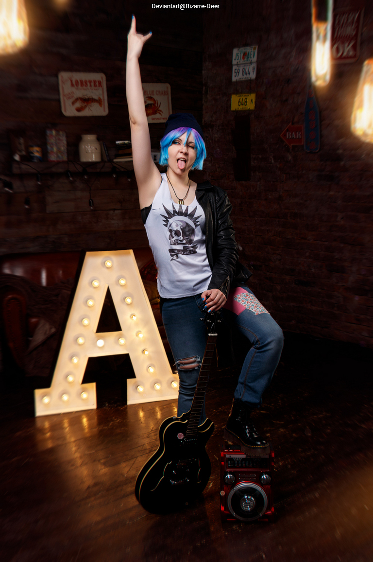 A young person with blue hair and punk attire poses with a guitar and makes a rock gesture, standing beside a large illuminated letter 'A' in a cozy setting.