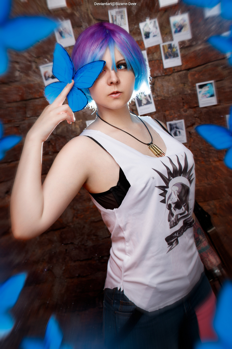 A person with short purple and blue hair poses with a large blue butterfly near their face, wearing a sleeveless top with a graphic design. The background features a brick wall with photographs.