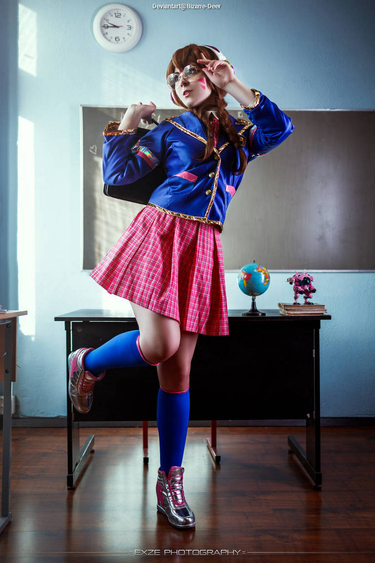 A person dressed in a blue jacket and plaid skirt poses playfully in a classroom setting, with a globe and clock visible in the background.