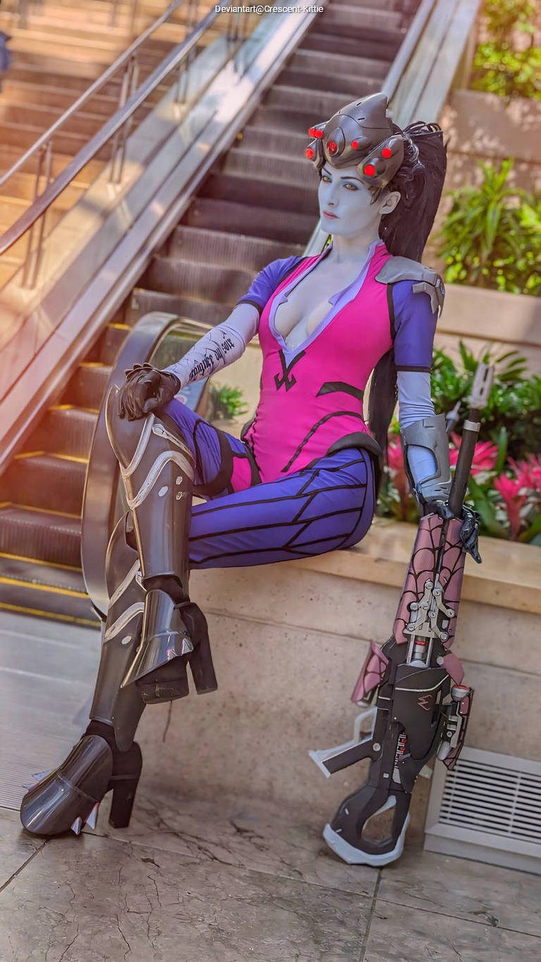 A person in a vibrant pink and blue cosplay outfit, resembling a futuristic character, sits on a ledge with an elaborate weapon, surrounded by greenery and escalators in the background.