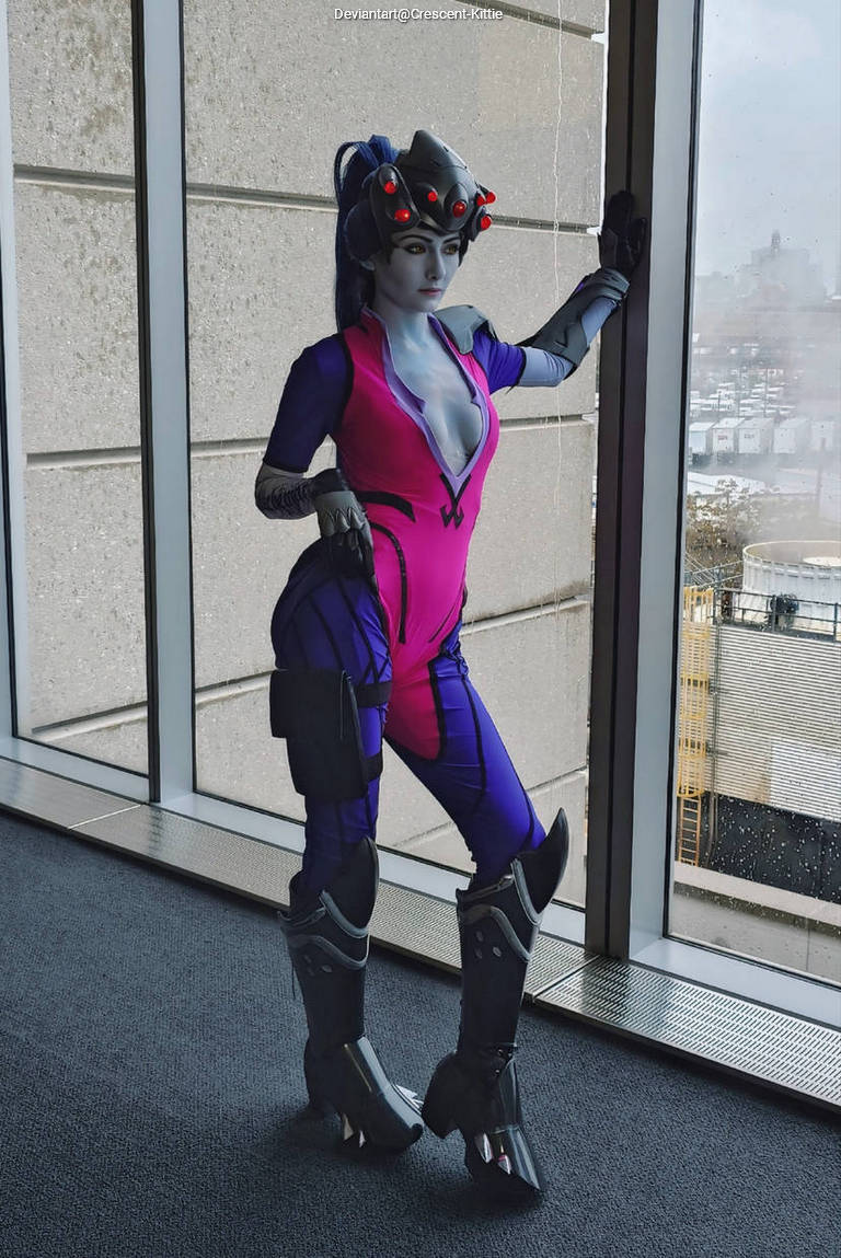 A cosplayer dressed as a character with a predominantly pink and purple outfit, featuring stylized armor and gloves, poses against a window with a cityscape visible outside.