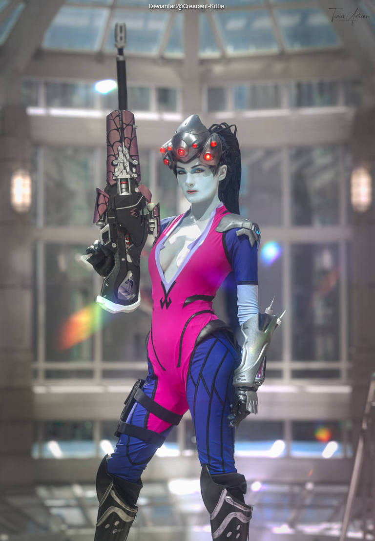 A person in a colorful, form-fitting costume holding a futuristic weapon, posing in a bright, modern environment.