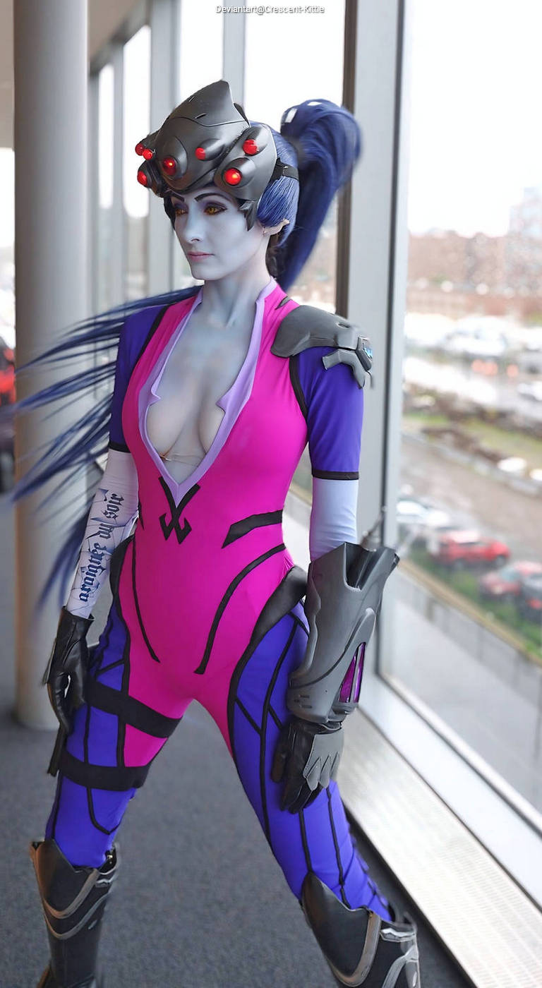 A person in a colorful cosplay outfit inspired by a character, featuring a tight pink and purple bodysuit, with futuristic armor elements and an elaborate headpiece, posing near a window.