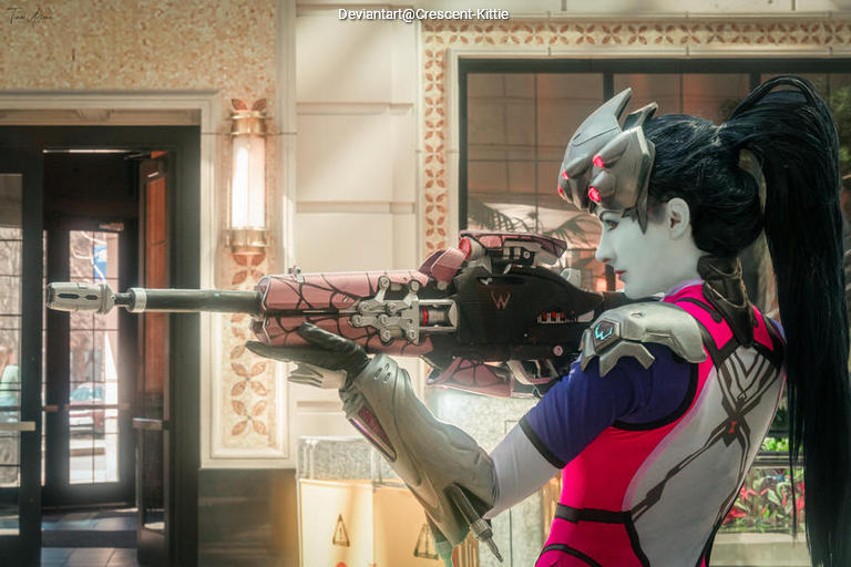 A character in a colorful costume holding a futuristic rifle, standing in a stylishly decorated interior space.