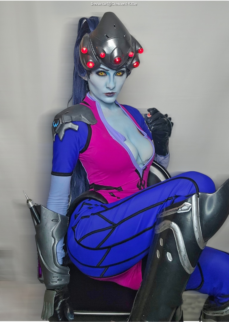 A person in a costume resembling a character with blue skin, wearing a pink and blue outfit, and a futuristic helmet with lights, sitting on a chair.
