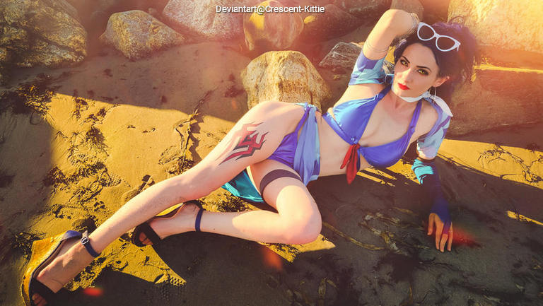 A person in a blue bikini lounging on a sandy beach, posing playfully among rocks, with sunglasses and a vibrant sunset in the background.