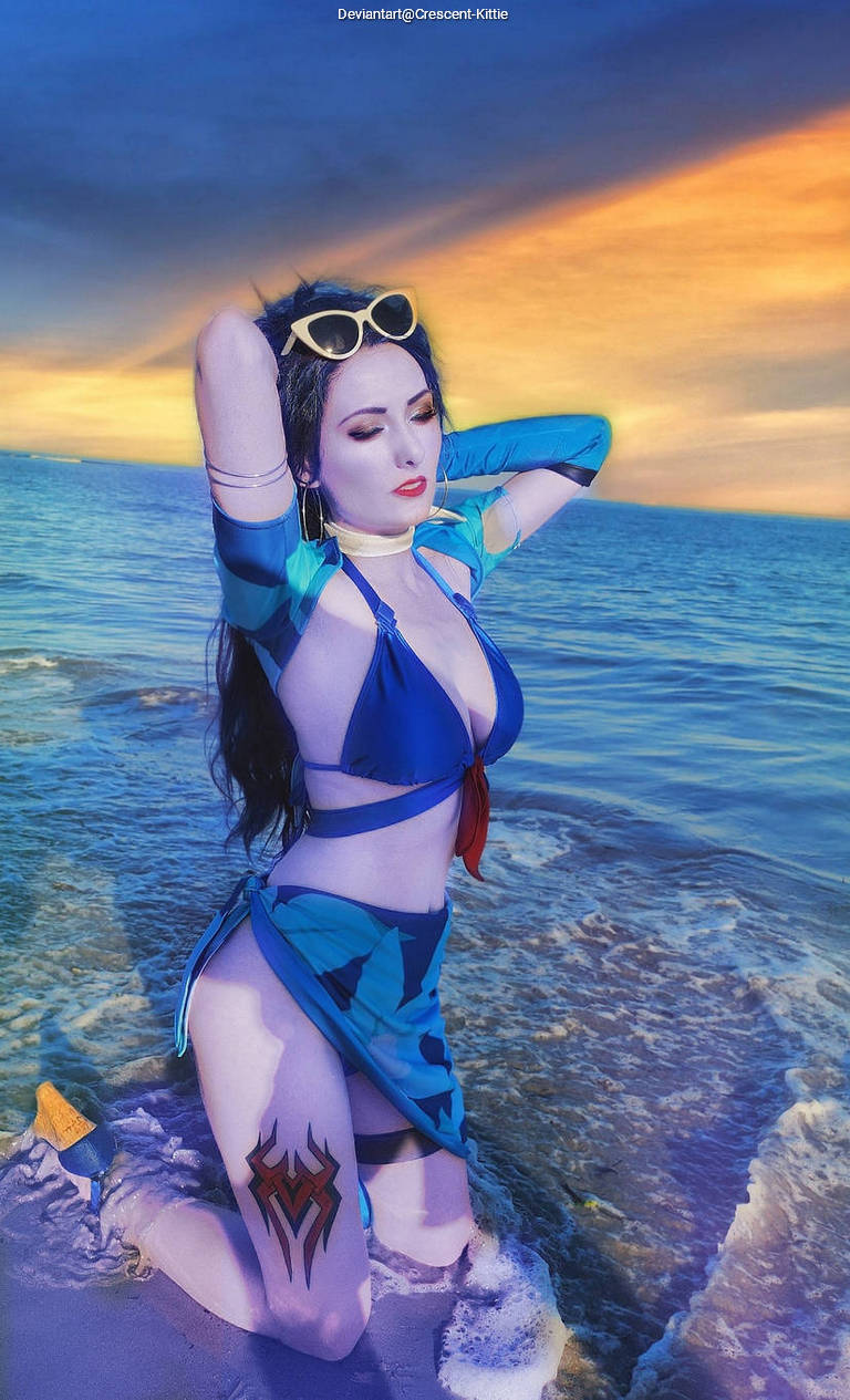 A woman in a blue bikini and matching cover-up kneels at the edge of the ocean, with waves lapping at her legs. She has long black hair, sunglasses, and a tattoo visible on her thigh. The background features a colorful sunset sky.