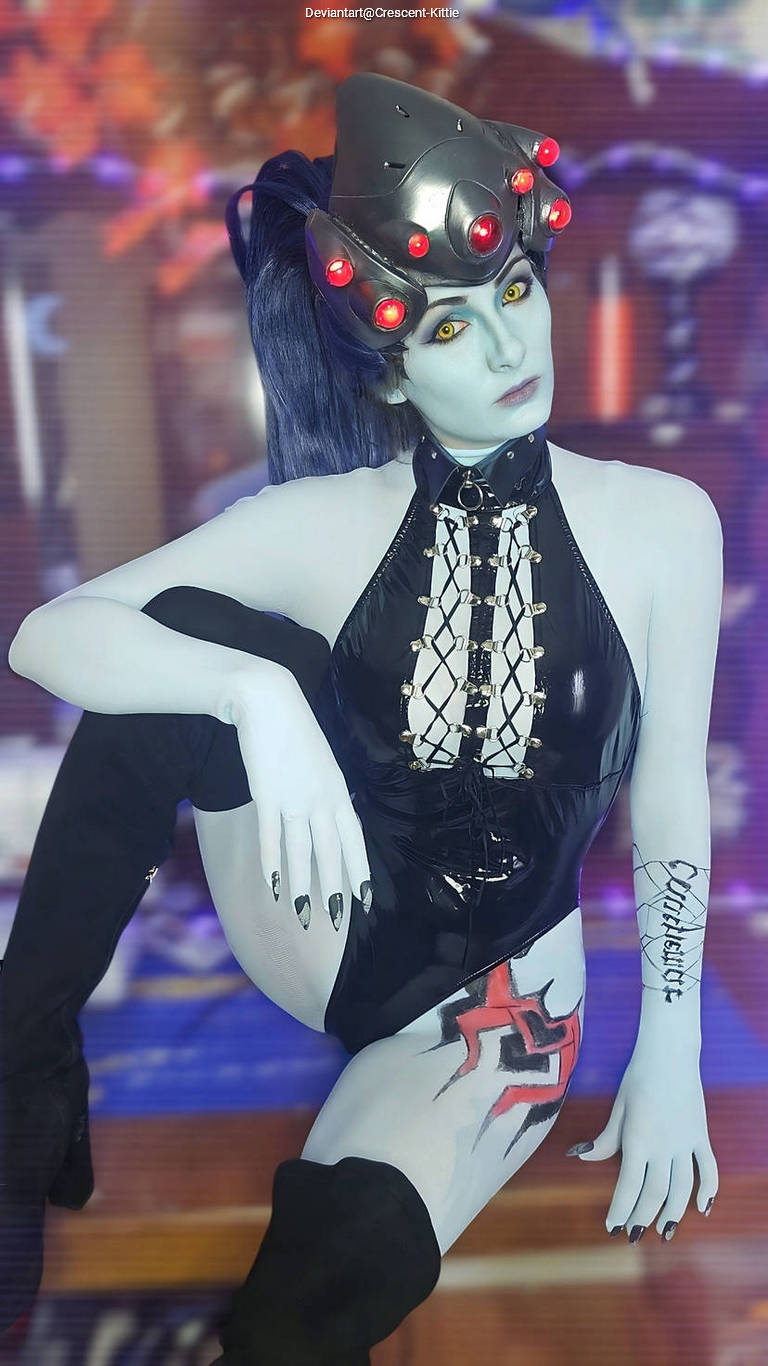 A person in a black latex outfit with a futuristic headpiece and bright yellow eyes poses dramatically, showcasing detailed body paint and a tattoo on their arm.