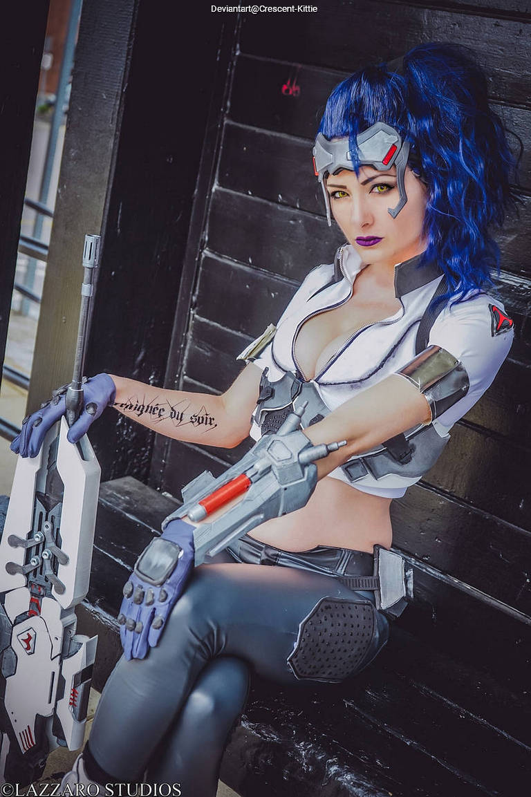 A female cosplayer dressed as a character with blue hair and futuristic attire, holding a mechanical weapon, posed in an urban setting.
