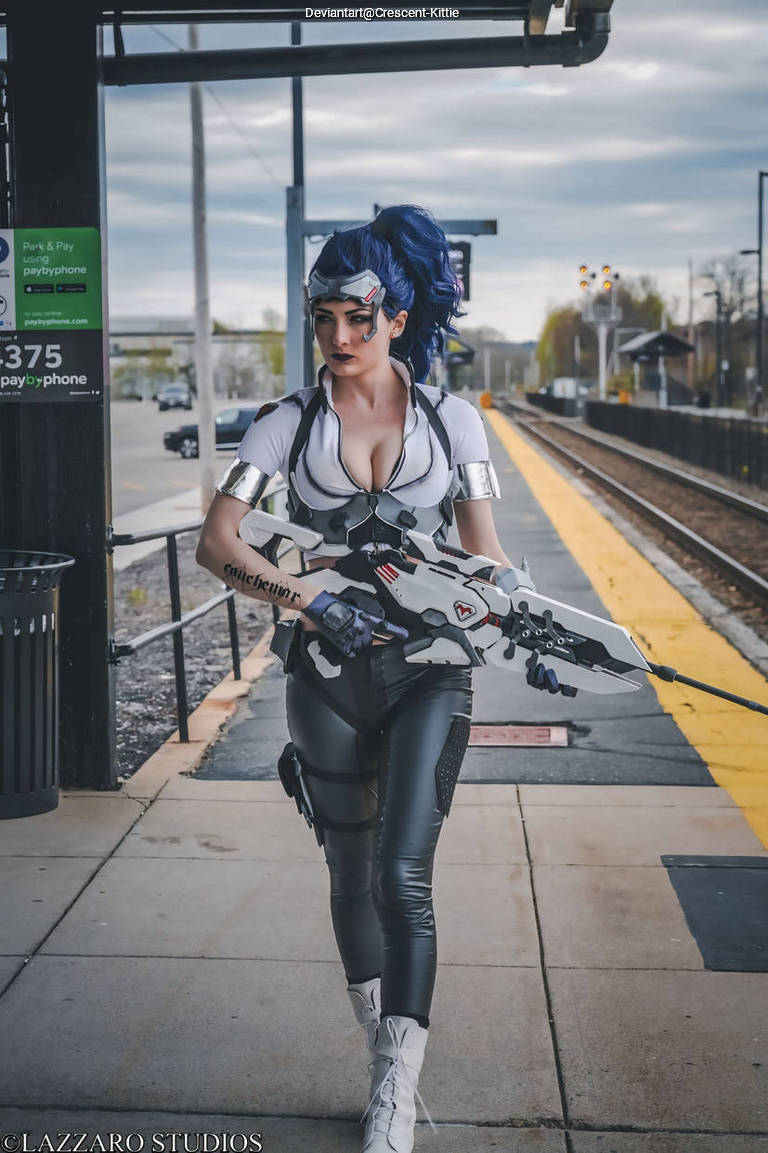 A person in a futuristic costume resembling a video game character, holding a large weapon, walking along a train platform.