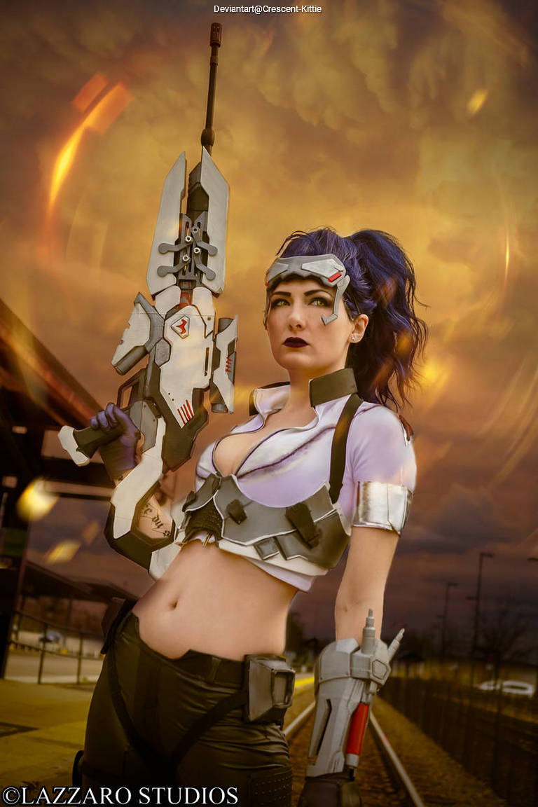 A female cosplayer dressed as a futuristic character holding a large weapon, set against a dramatic sky and train tracks.