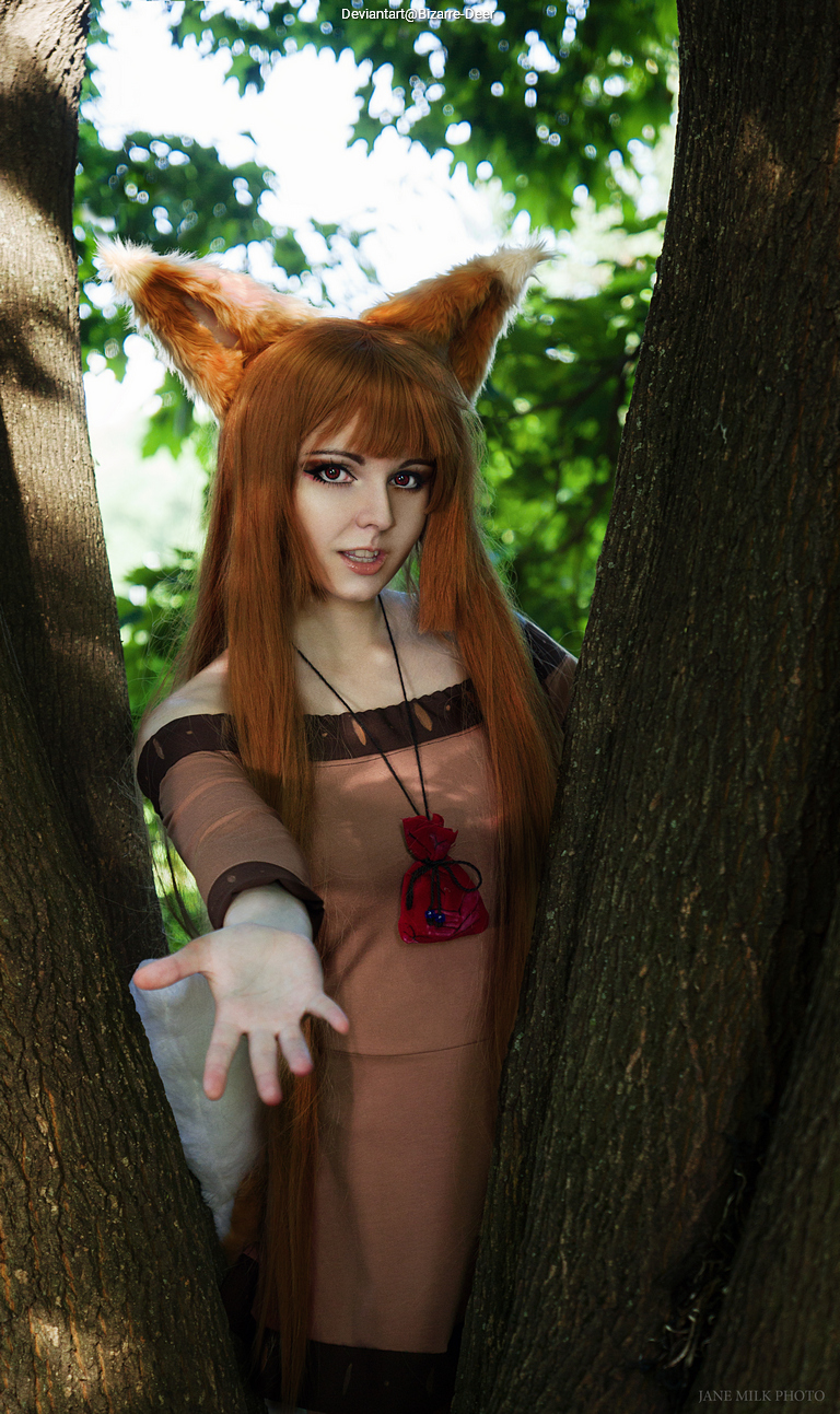 A person with fox-like ears and long orange hair is reaching out with one hand between two tree trunks, wearing a brown dress and a red pouch around their neck.