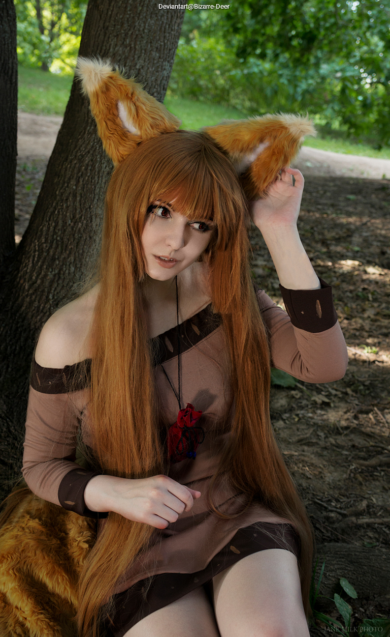 Cosplayer with long brown hair and fox ears sitting against a tree in a natural setting, wearing a brown off-shoulder dress.