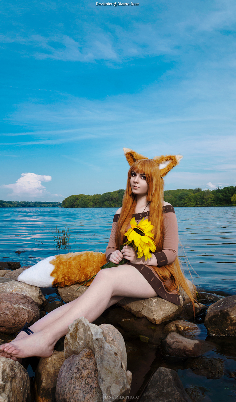 A person in a fox costume with orange and white ears and a tail sits on rocks by a body of water, holding a sunflower. The background features a blue sky with clouds and green trees.