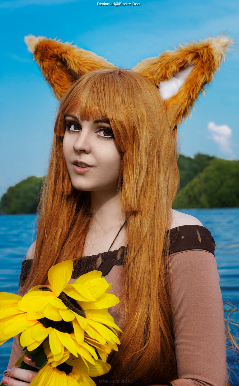 A person with long, orange hair and fox ears holding a bouquet of sunflowers, standing by a body of water with greenery in the background.