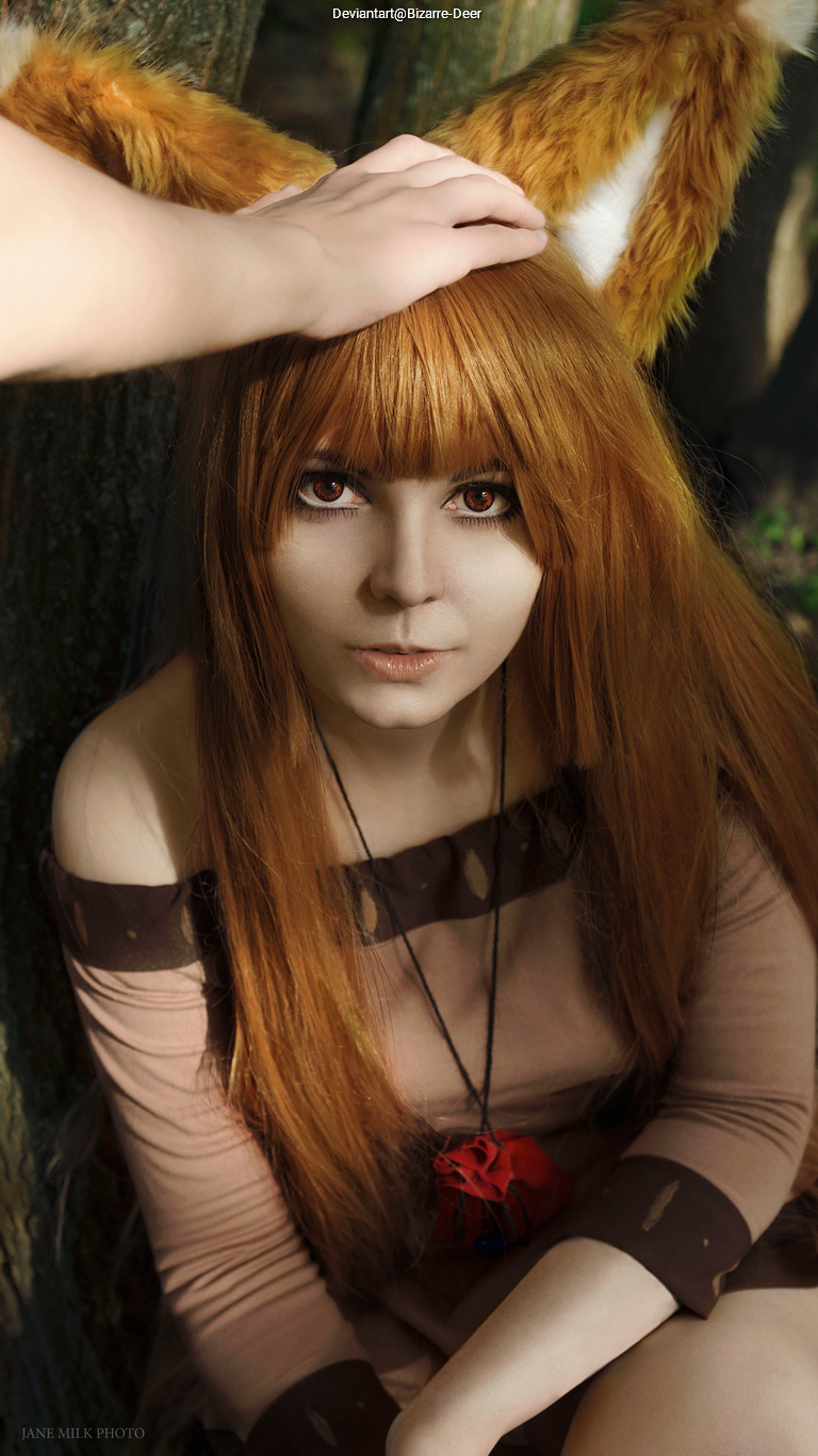 A person with long, orange fox ears and brown hair, wearing a brown outfit, sitting against a tree with a hand gently resting on their head.