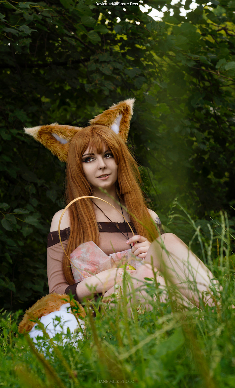 A person with long brown hair and fox ears sitting in a grassy area, holding a plush toy and wearing a light brown outfit with off-the-shoulder sleeves.