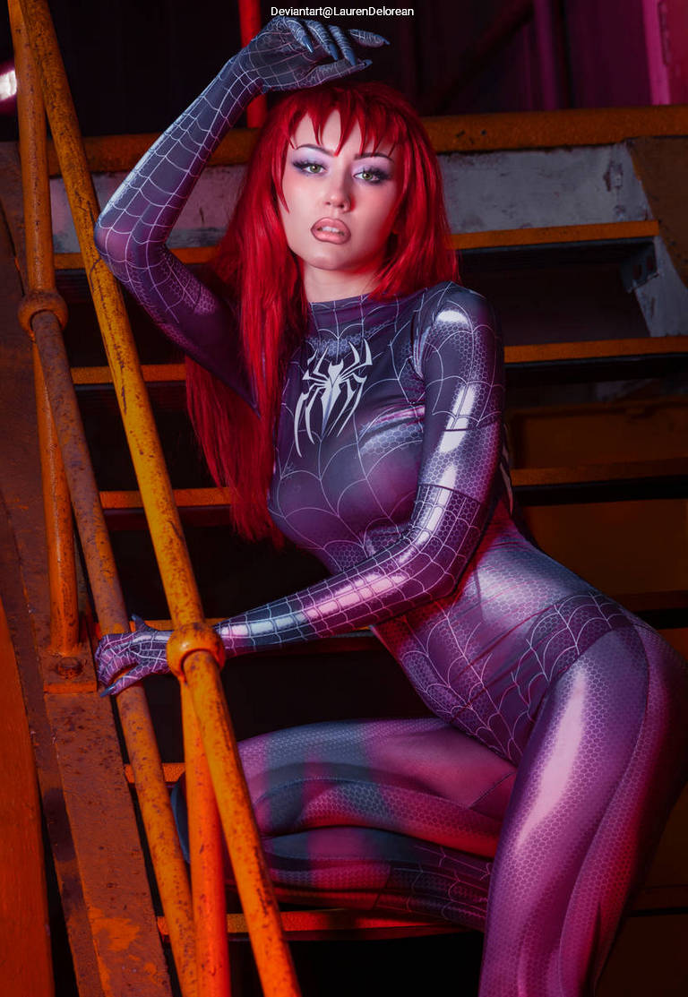 A person with long red hair poses confidently on a metal staircase, wearing a form-fitting, shiny spider-themed costume. The background features industrial elements with a mix of colors, creating a vibrant atmosphere.