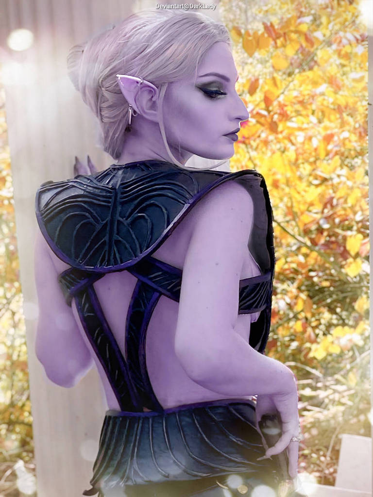 A person with pale purple skin and pointed ears wearing a black and purple costume, viewed from behind with a natural autumn background.