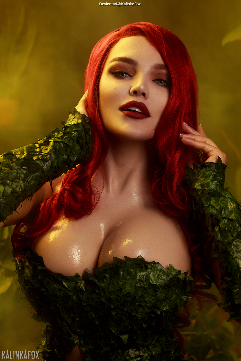 A person with long red hair styled in a glamorous way, wearing a costume adorned with green leaves, posing confidently against a soft, warm background.