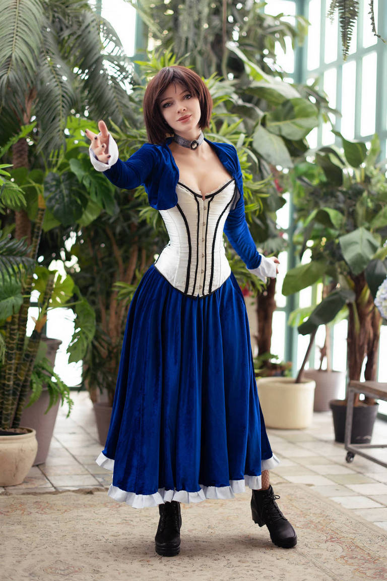 A person in a blue dress with a corset-style top is posing confidently in a botanical setting, surrounded by lush green plants.