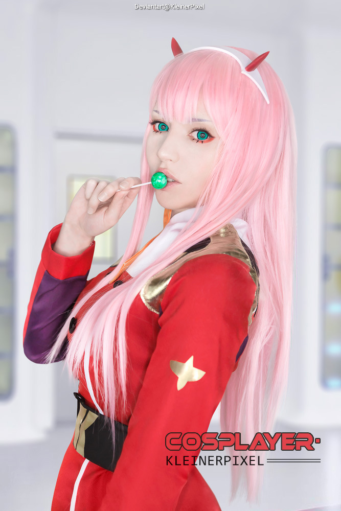 A cosplayer in a red outfit with gold and purple accents, featuring long pink hair and green eyes, holding a green lollipop.