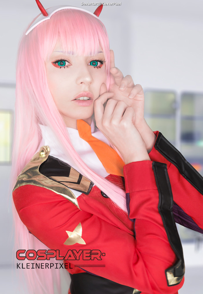 Portrait of a cosplayer with long pink hair and blue-green eyes, wearing a red and black costume with a white collar and orange accents, posing with a subtle expression in a bright background.