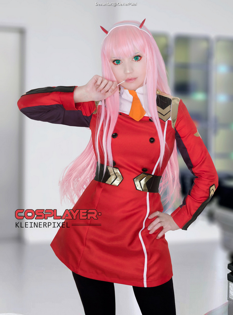 A person in a red cosplay outfit with a pink wig and green eyes, posing confidently with one hand on their hip and a lollipop in the other.
