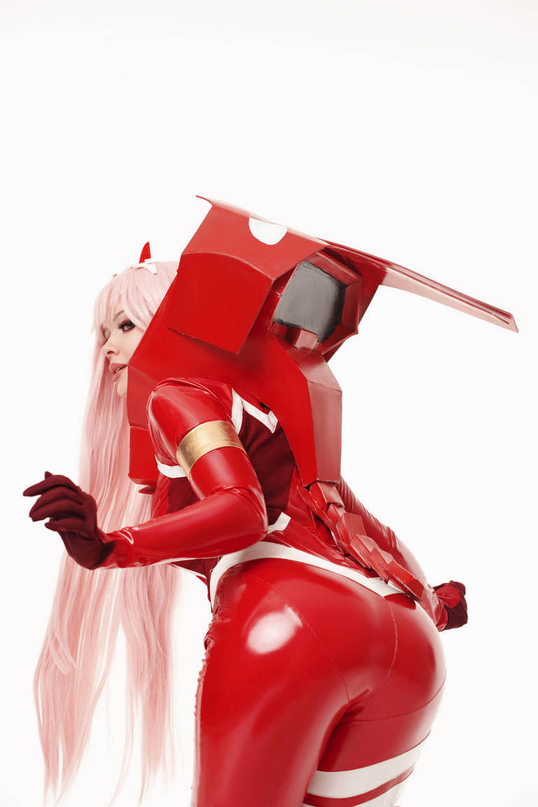 Cosplayer wearing a shiny red outfit with a large mechanical accessory on the back, posed dynamically against a white background.