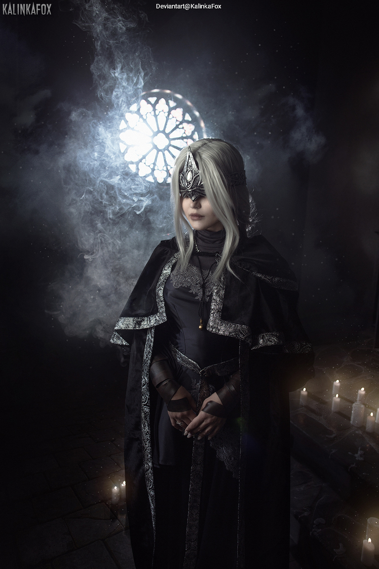 A person dressed in a dark, ornate costume with a crown-like headpiece, standing in a dimly lit setting with fog and candles. A rose window is visible in the background.