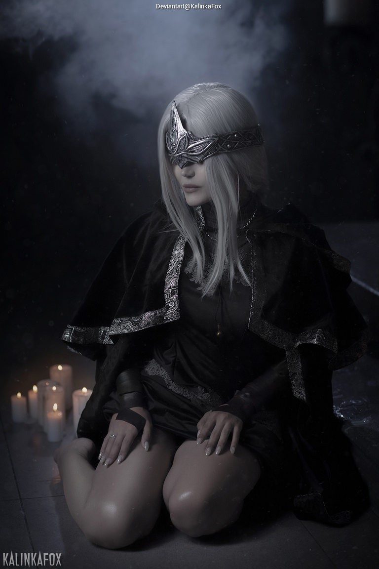 A character in dark clothing with long white hair kneels on the floor surrounded by candles, wearing a decorative headpiece and a veil over their eyes.