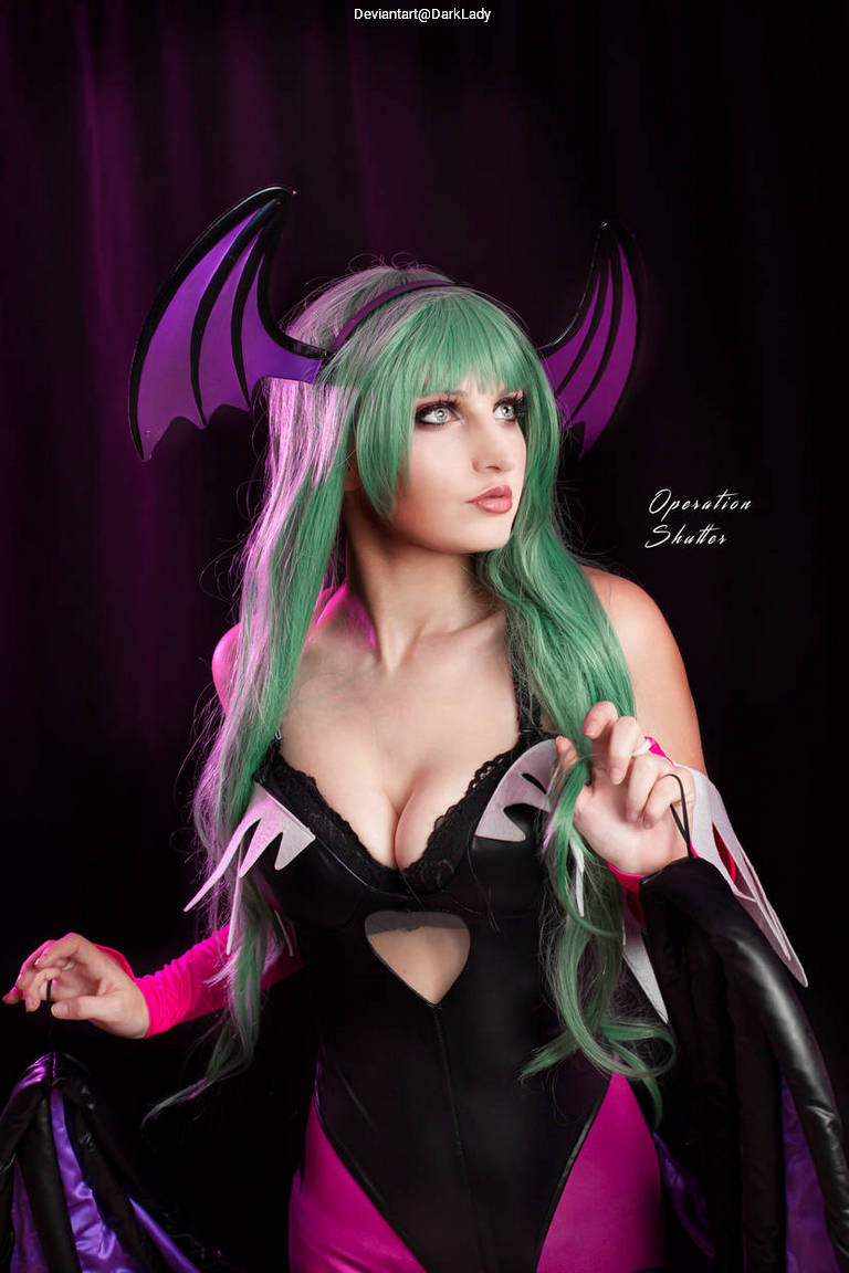 A model dressed in a black and pink costume with bat-like wings and green hair striking a pose against a dark purple background.