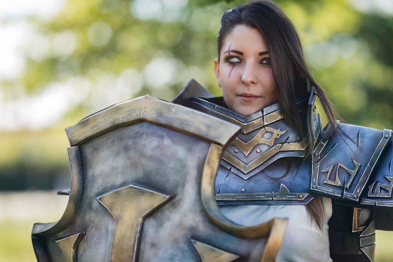 A woman in detailed armor and a large shield, with striking makeup and a serious expression, set against a blurred outdoor background.
