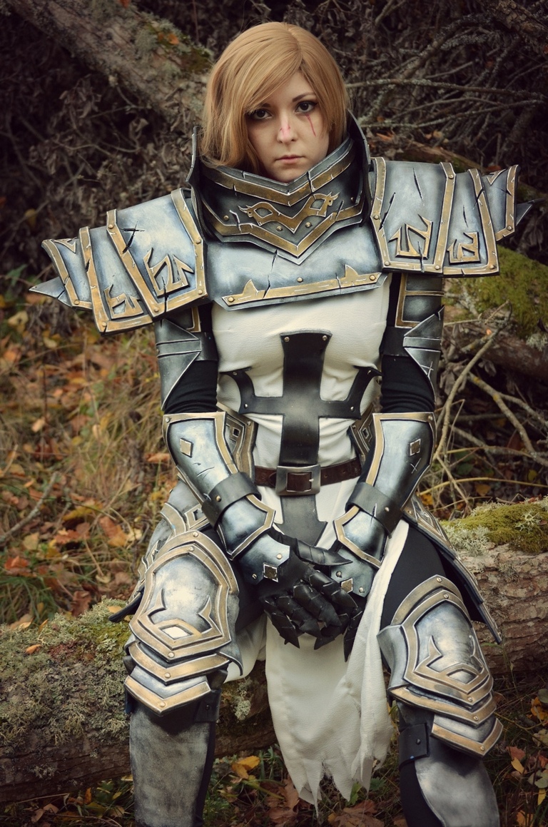 A person dressed in intricate silver and black armor, seated on a log in a forest setting, with a serious expression.