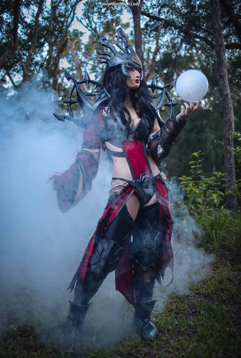 A female cosplayer dressed in a dark, elaborate costume with a crown and armor, standing in a forest with mist surrounding her while holding a glowing orb.