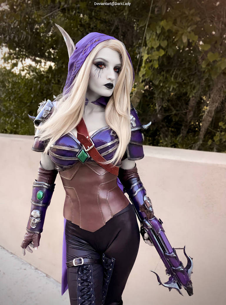 A person in a detailed fantasy costume with a purple cape, hood, and armor, portraying a character with pale skin and striking features, set against a natural background.