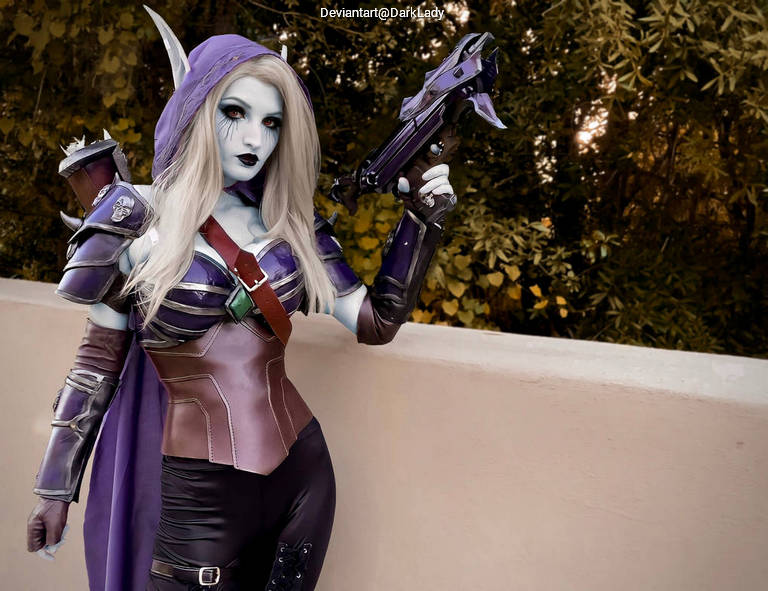 A cosplayer dressed in a fantasy-themed outfit featuring a purple cape and armor, holding a weapon, with an outdoor background.