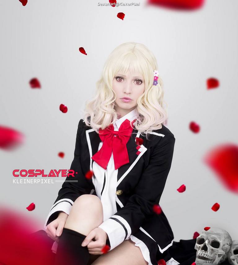 A cosplayer with long, wavy blonde hair wearing a black school uniform with a red bow tie, sitting among falling rose petals and a decorative skull.