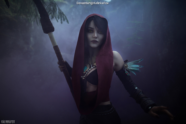 A character dressed in a dark hooded outfit stands in a misty forest, holding a staff in one hand. The scene conveys a mystical atmosphere.