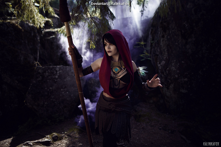A character in a forest setting, wearing a red hood and a decorative outfit, holding a staff and appearing contemplative against a background of mist and rocks.