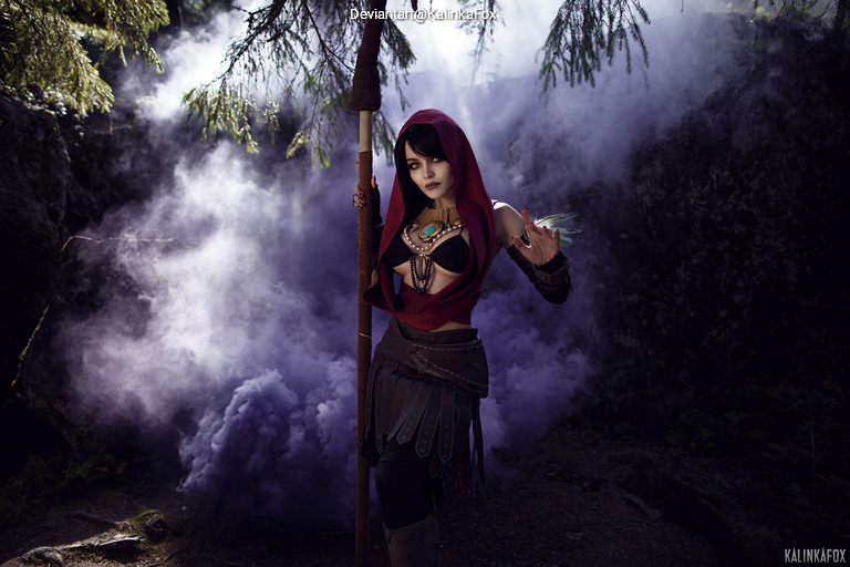 A cosplayer dressed as a mystical character stands amidst purple smoke, holding a staff and adorned with intricate jewelry, surrounded by trees.