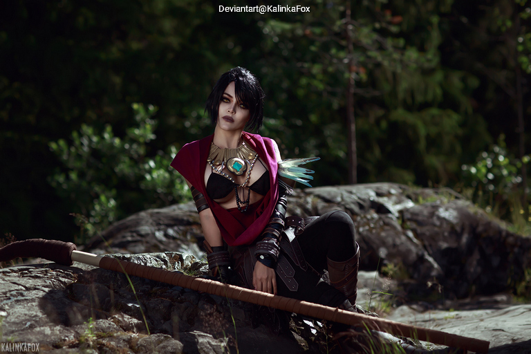 A person in a dark outfit with a red cape and decorative accessories, seated on a rock in a forest setting, exuding a mysterious aura.